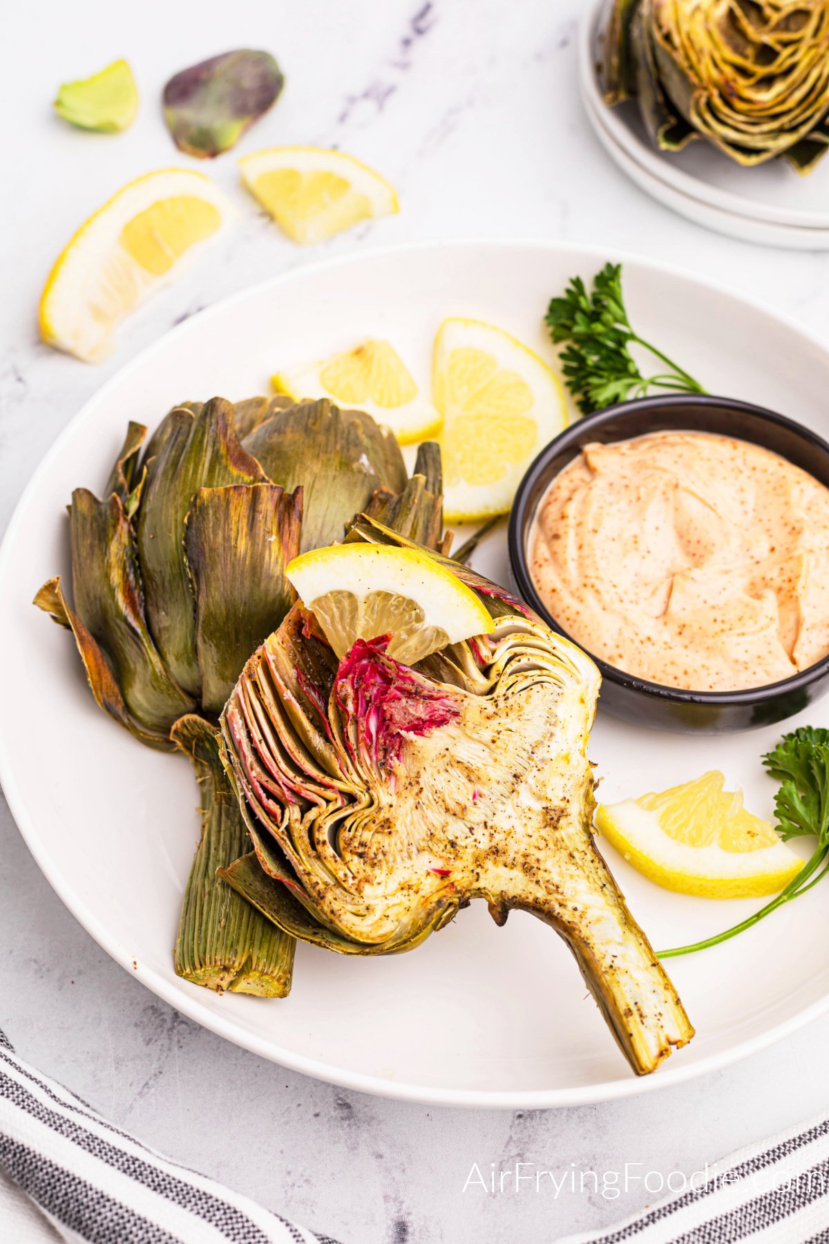Air fried artichoke on a white plate with a side of lemons and dipping sauce.