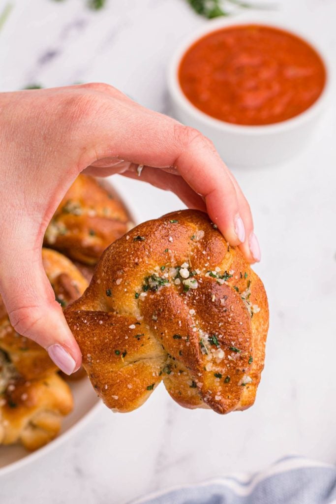 Hand holding golden garlic knot with small bowl of red sauce on the table