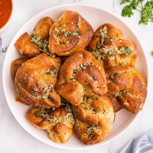 Golden bread knots with garlic and parmesan cheese stacked on a white plate with a small bowl of red sauce.