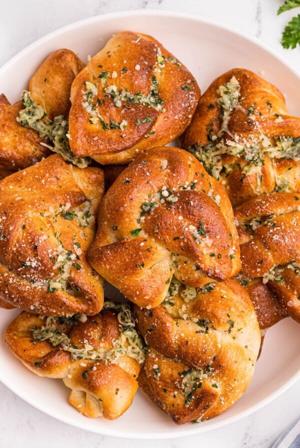 Golden bread knots with garlic and parmesan cheese stacked on a white plate with a small bowl of red sauce.