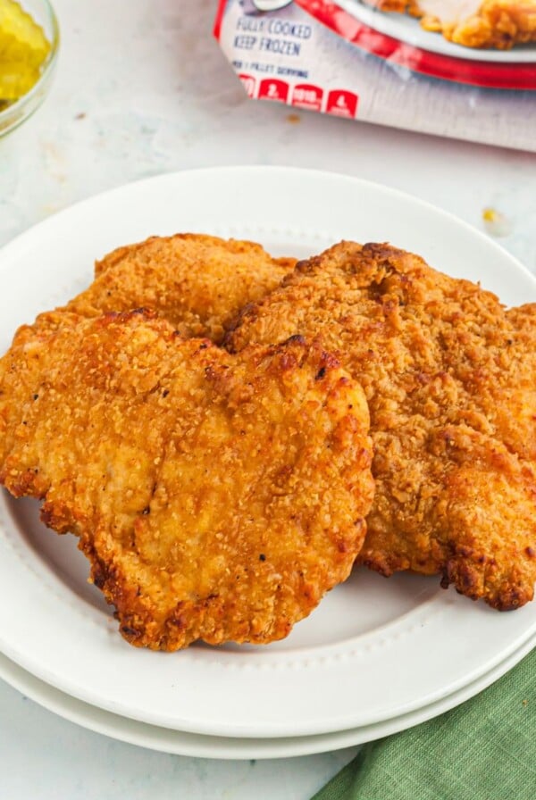 Golden crispy chicken breasts on a white plate in front of pickles and a red bag of chicken