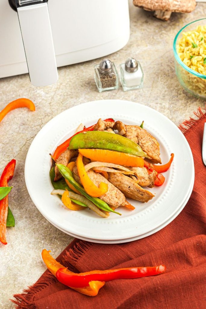 Chicken stir fry with peppers and mushrooms on a white plate in front of an air fryer and salt and pepper shakers.