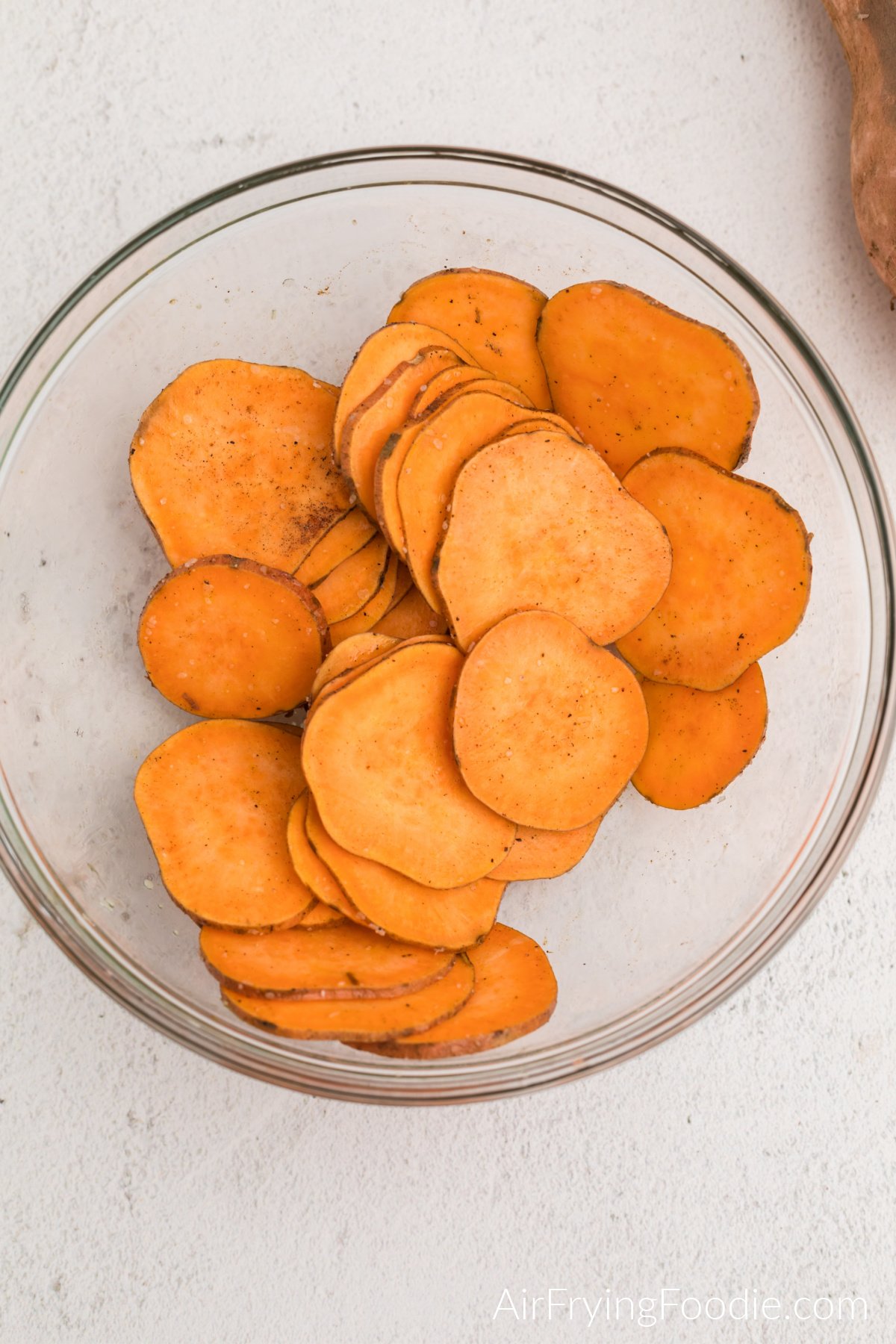 Sliced sweet potatoes in a glass bowl coated with olive oil and seasonings.