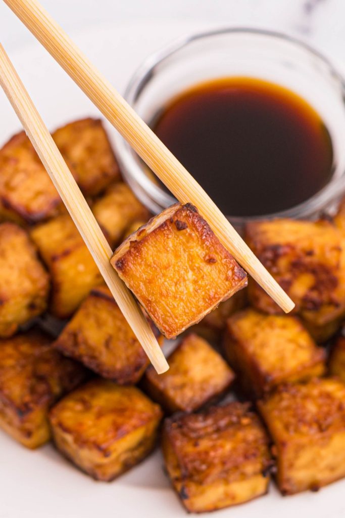 Golden crispy tofu held between chopsticks over a plate of tofu cubes