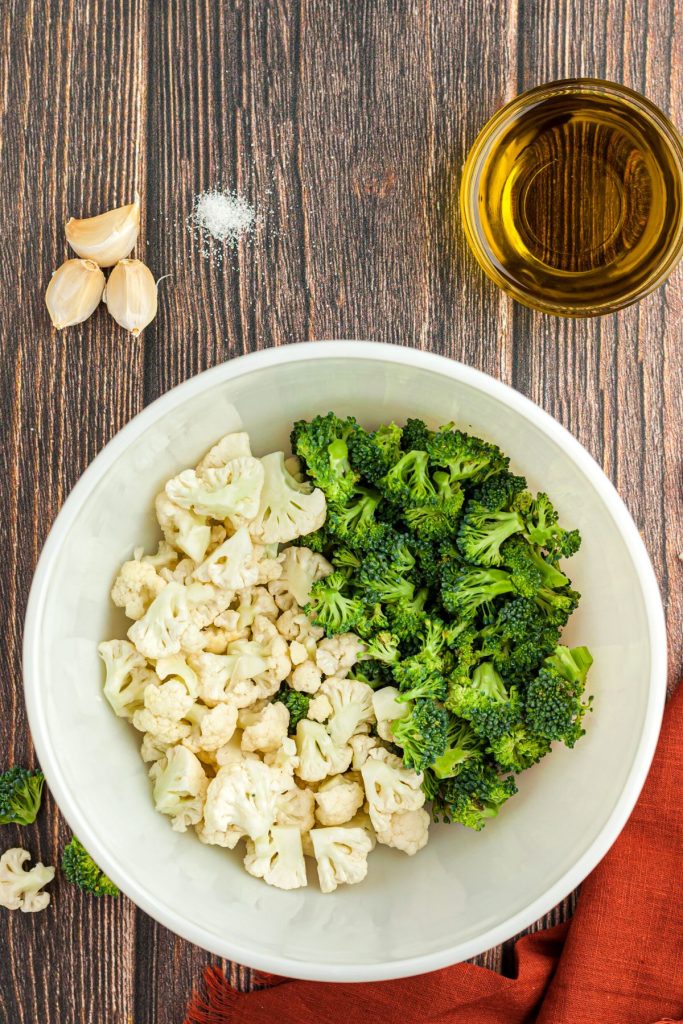 Broccoli and cauliflower florets in a white bowl with garlic cloves, salt, and olive oil. 