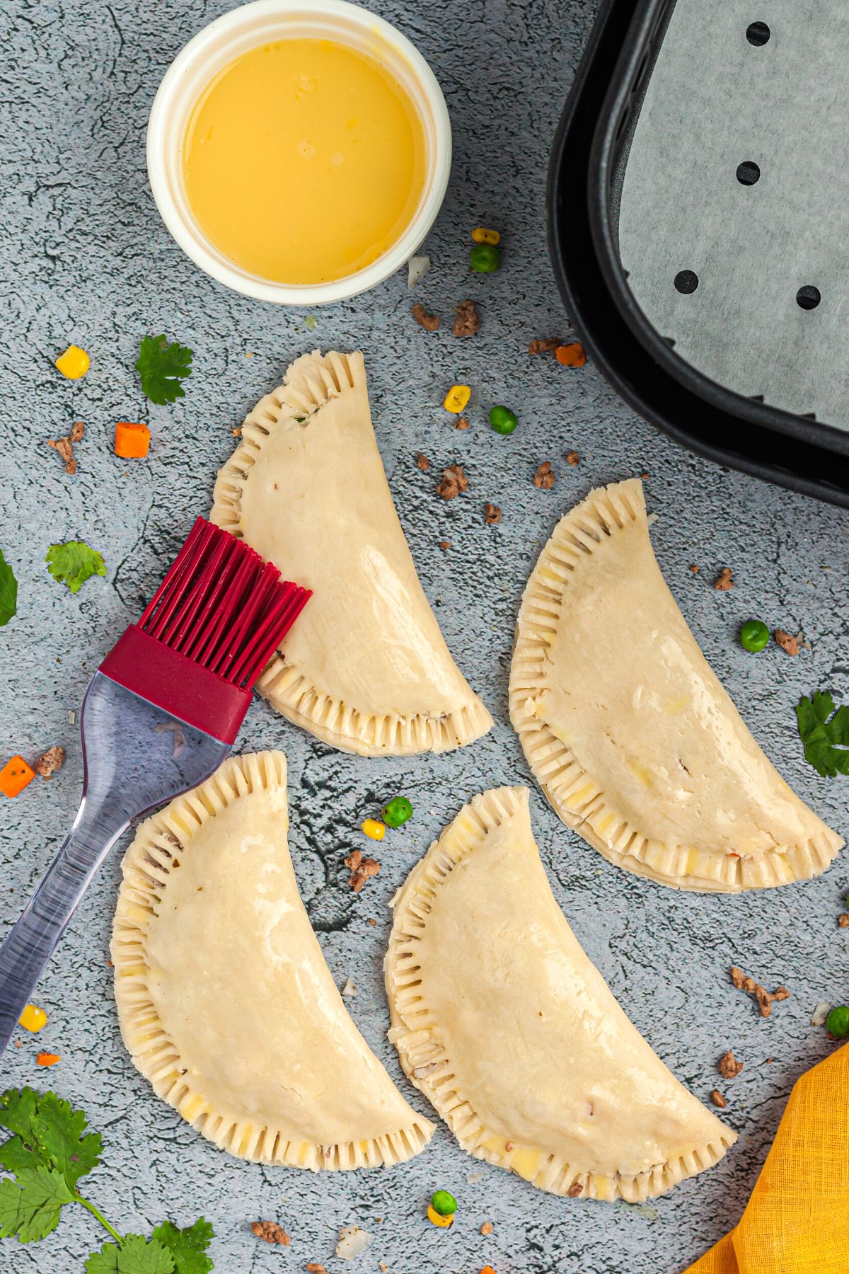 Uncooked empanadas on a light blue table in front of air fryer basket being brushed with egg wash