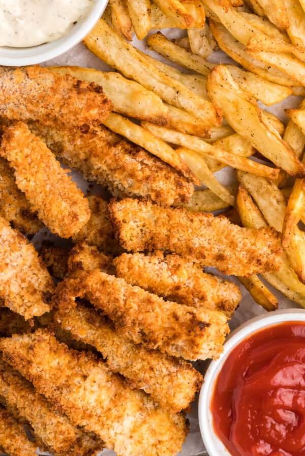 Fish and chips on a baking sheet with ketchup and dipping sauce.