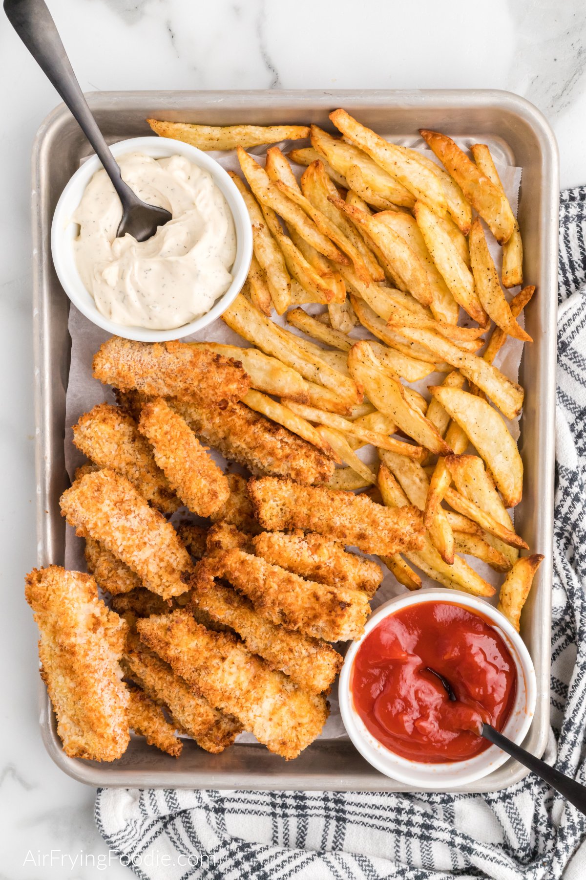 Air fryer fish and chips on a baking sheet with ketchup and dipping sauce.