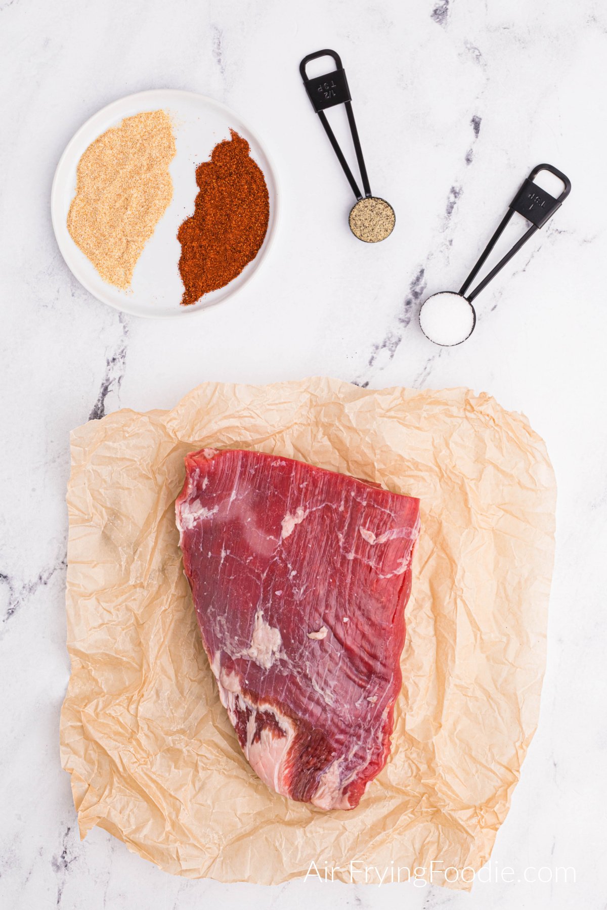 Flank steak on butcher paper and seasonings in a bowl and in measuring spoons on a white table. 