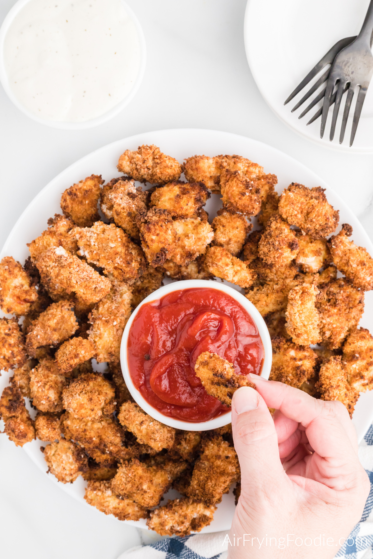 Popcorn chicken on a serving plate, with a hand dipping a pice of chicken into a bowl of ketchup.