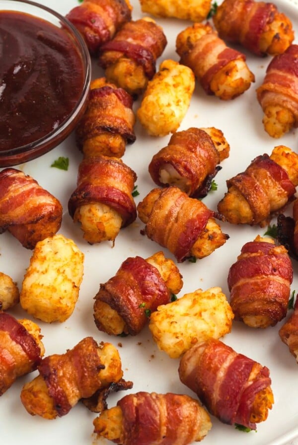 Golden and crispy bacon wrapped tater tots on a white round plate served with bbq sauce