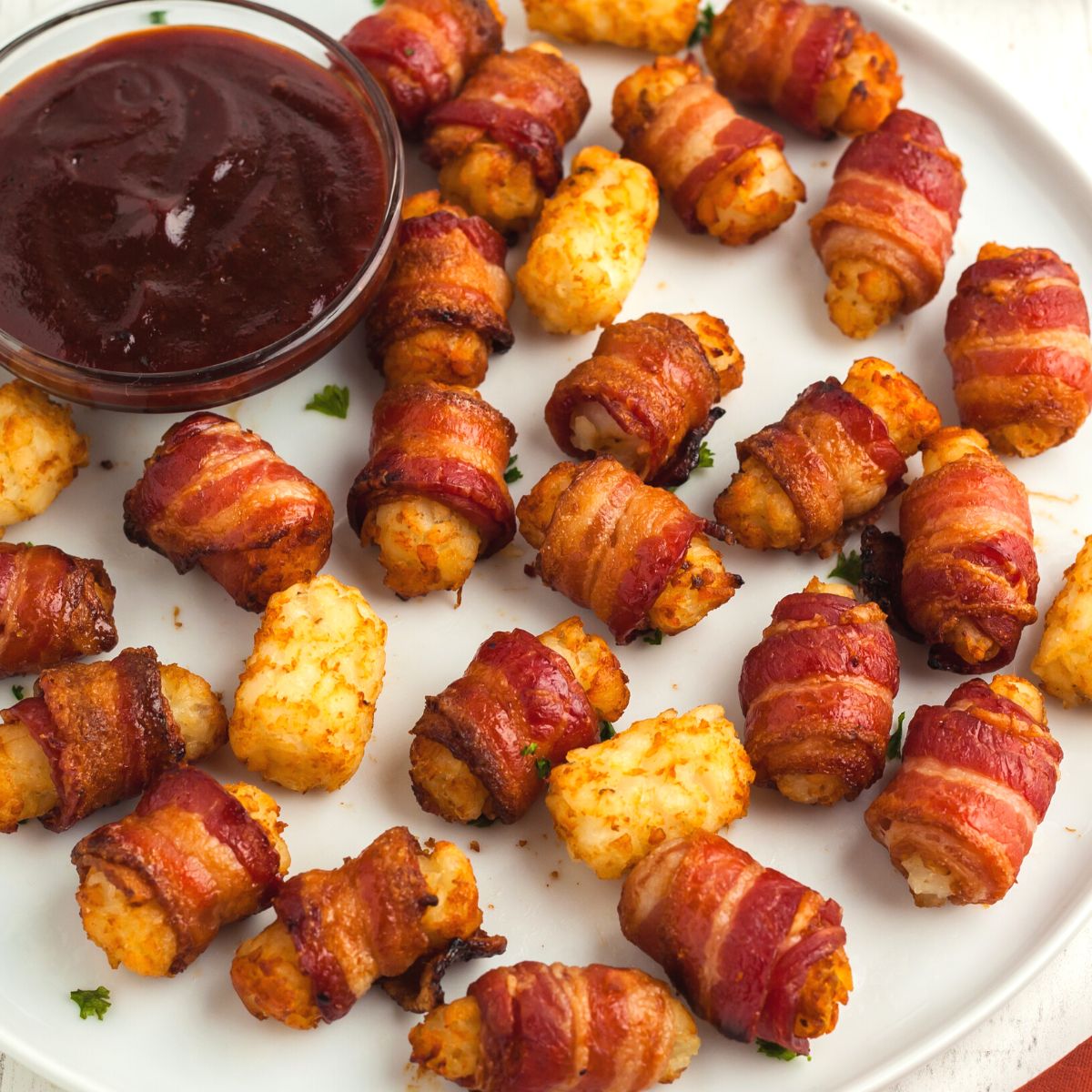 Golden and crispy bacon wrapped tater tots on a white round plate served with bbq sauce