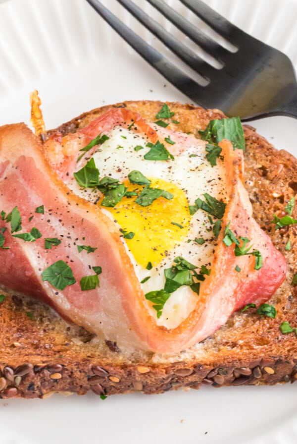 Fully cooked air fryer toast with bacon and egg served on a round white plate with a fork.