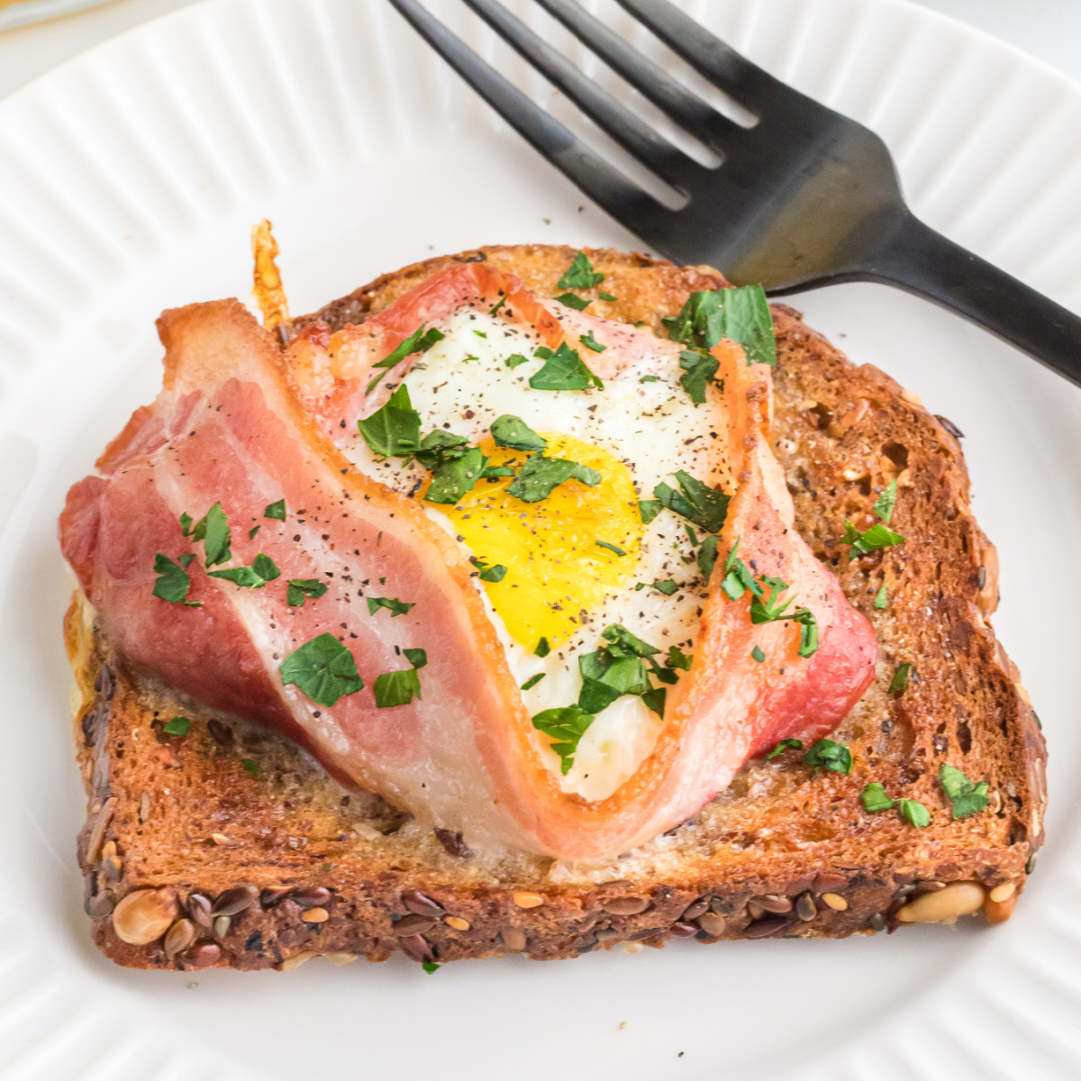 Fully cooked air fryer toast with bacon and egg served on a round white plate with a fork.