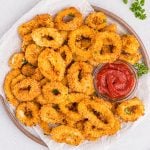 Close-up of golden brown calamari cooked in the air fryer served on a round white plate with a small glass bowl of cocktail sauce on the plate to the right.