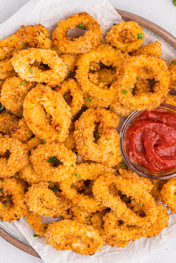 Close-up of golden brown calamari cooked in the air fryer served on a round white plate with a small glass bowl of cocktail sauce on the plate to the right.