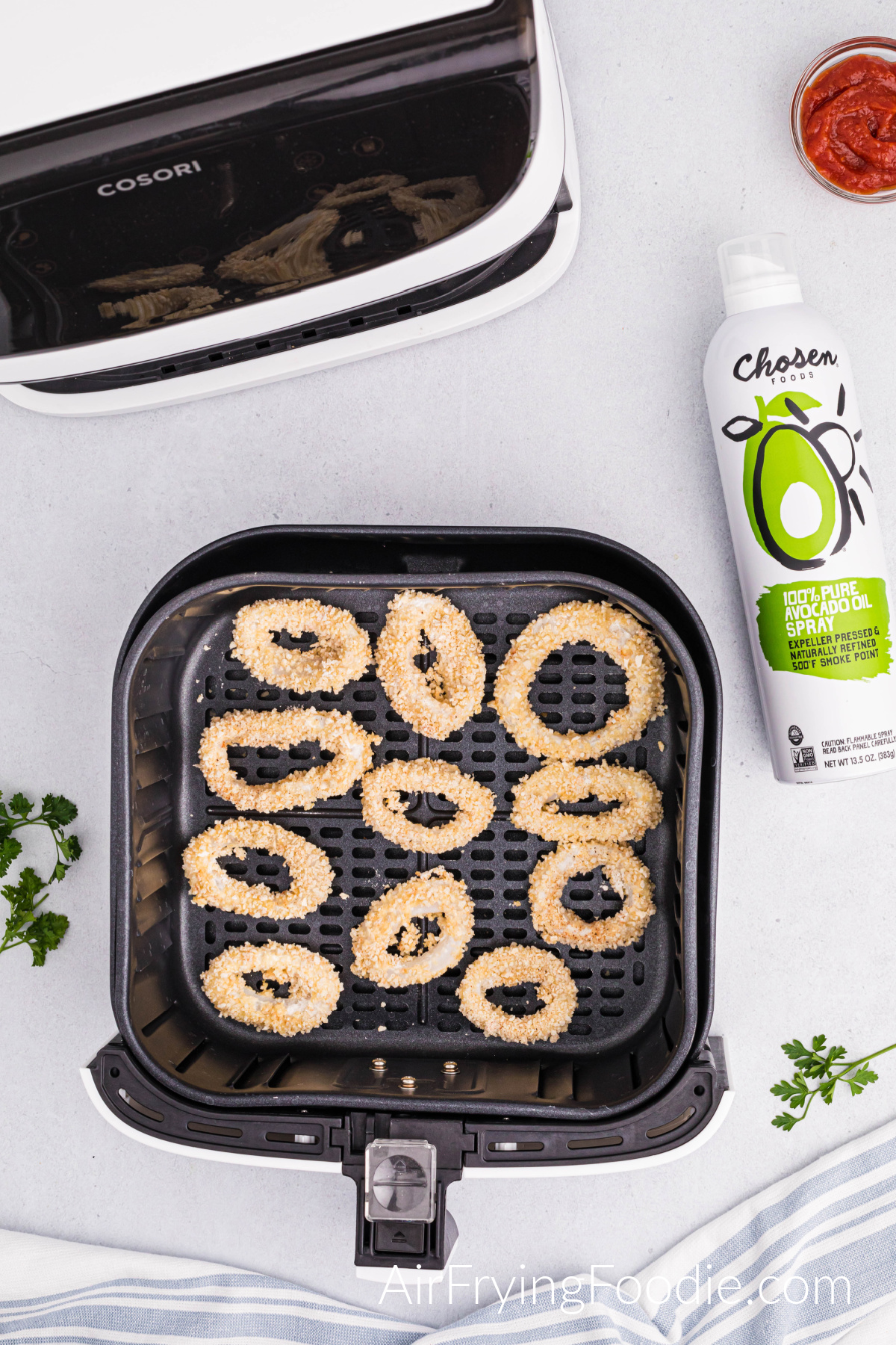 A picture of the air fryer basket with calamari, not overlapping, that has been dipped and ready to air fry. A small portion of the air fryer base is in the top left of the picture, and a can of avocado oil and a small glass bowl of cocktail sauce is to the top right.