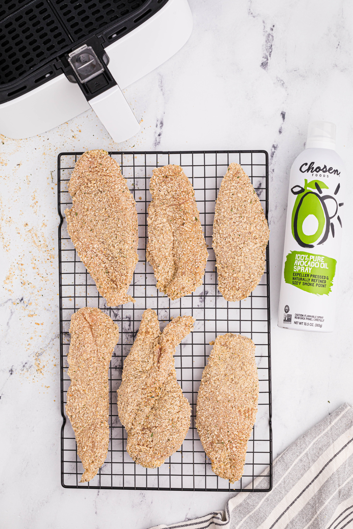Six chicken cutlets fully covered in breadcrumb mixture resting on a cooling rack before going in the air fryer. A can of olive oil spray is lying to the right of the cooling rack, with a small portion of the air fryer at the top of the picture.