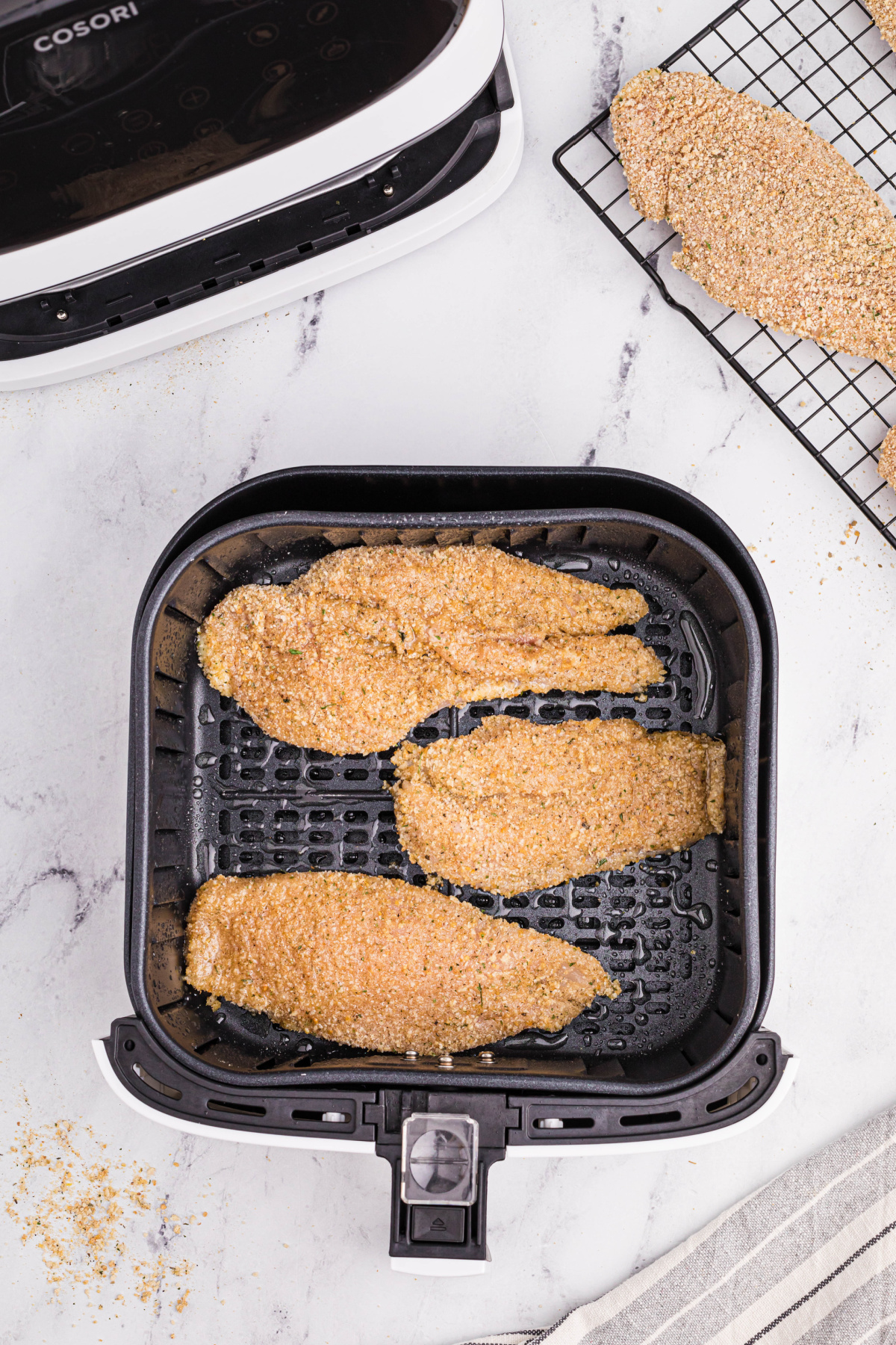 Three uncooked breaded chicken cutlets in the air fryer basket with a small portion of the cooling rack with a breaded chicken cutlet and the base of the air fryer are at the top of the picture.