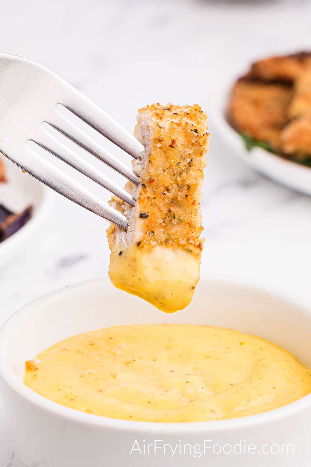 A white bowl of creamy honey mustard sauce with a piece of fully cooked chicken from the air fryer on a fork dipped in the creamy honey mustard sauce. 