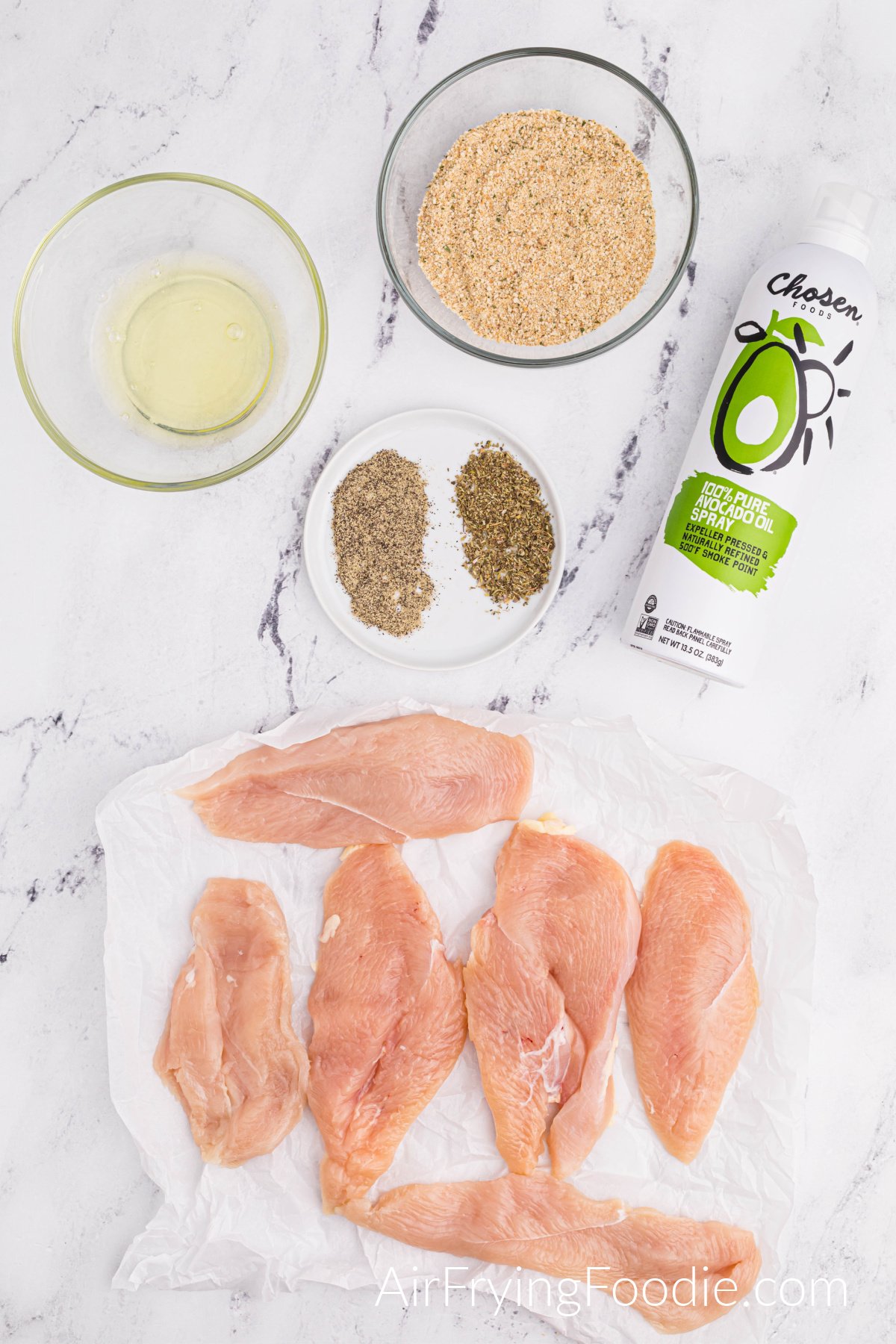 Ingredients picture with six boneless, skinless raw chicken breasts, a round white plate with Italian seasoning, and ground black pepper. Two clear bowls consist of egg whites, and the other contains seasoned breadcrumbs and a bottle of olive oil spray.