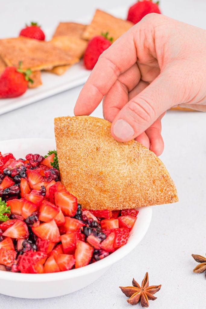 A hand lifting a cinnamon sugar tortilla chip out of a bowl of chopped fruit