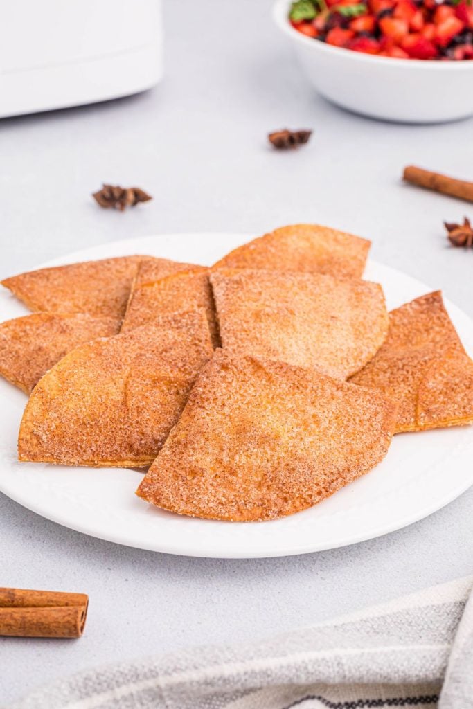 Sugar and cinnamon coated tortilla chips on a white plate in front of an air fryer and bowl of chopped strawberries