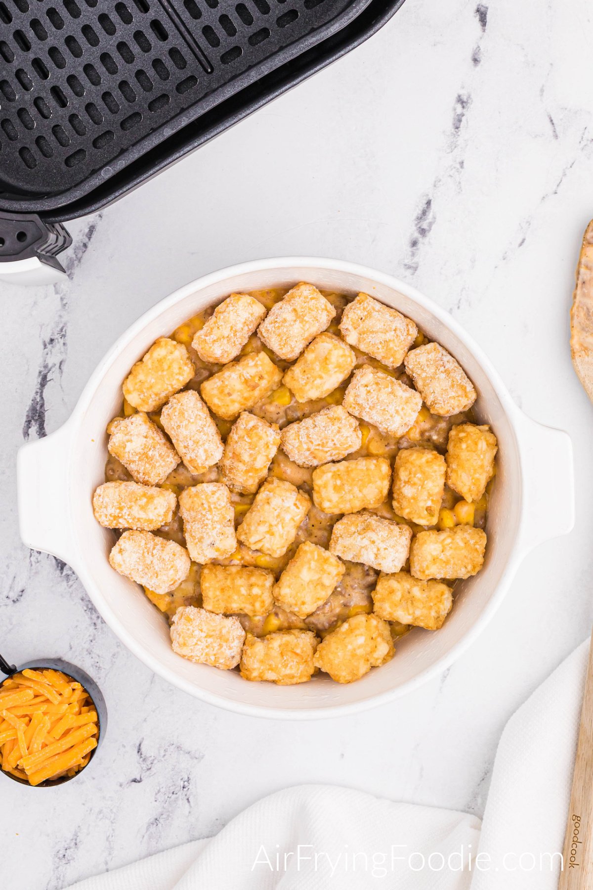 A round white bowl with layers of tater tots and seasoned ground beef with tater tots as the top layer. A small portion of the air fryer basket and wooden spoon are also in the picture, with a small measuring cup of shredded cheddar cheese.