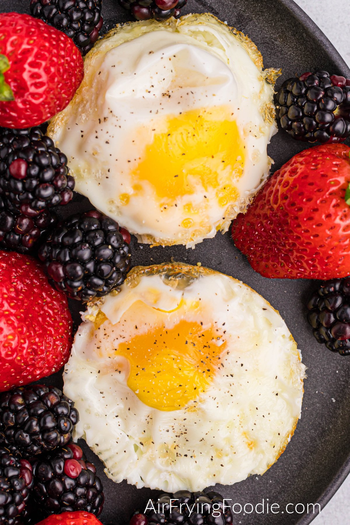 Air fryer fried eggs topped with salt and pepper and served on a plate with a side of fruit.
