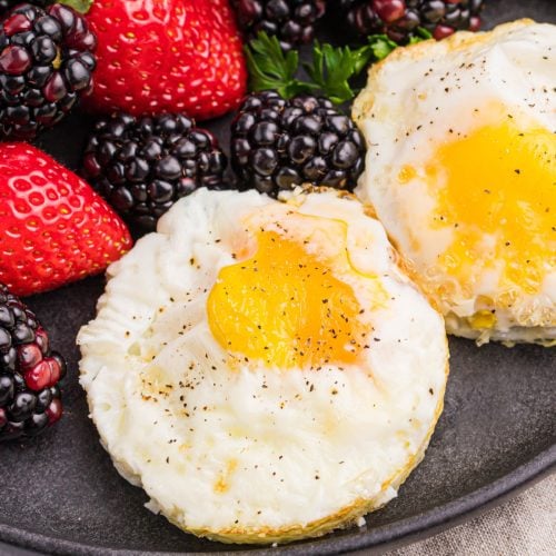 Air fryer fried eggs topped with salt and pepper and served on a dark plate with a side of fruit.