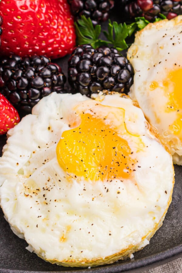 Air fryer fried eggs topped with salt and pepper and served on a dark plate with a side of fruit.