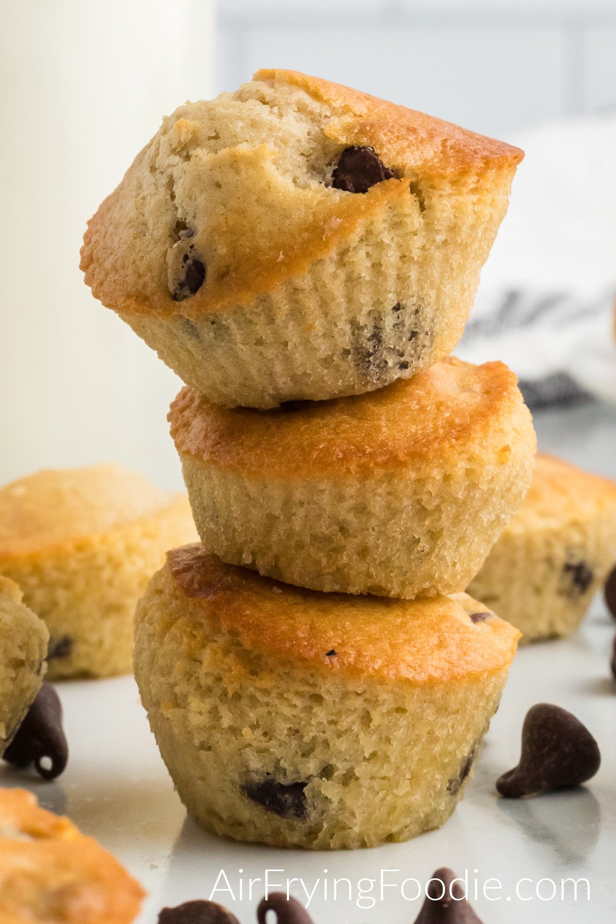 Stack of three air fryer chocolate chip muffins with more muffins beside and behind the stack and chocolate chips sprinkled in front of the stack of three muffins on the table. 
