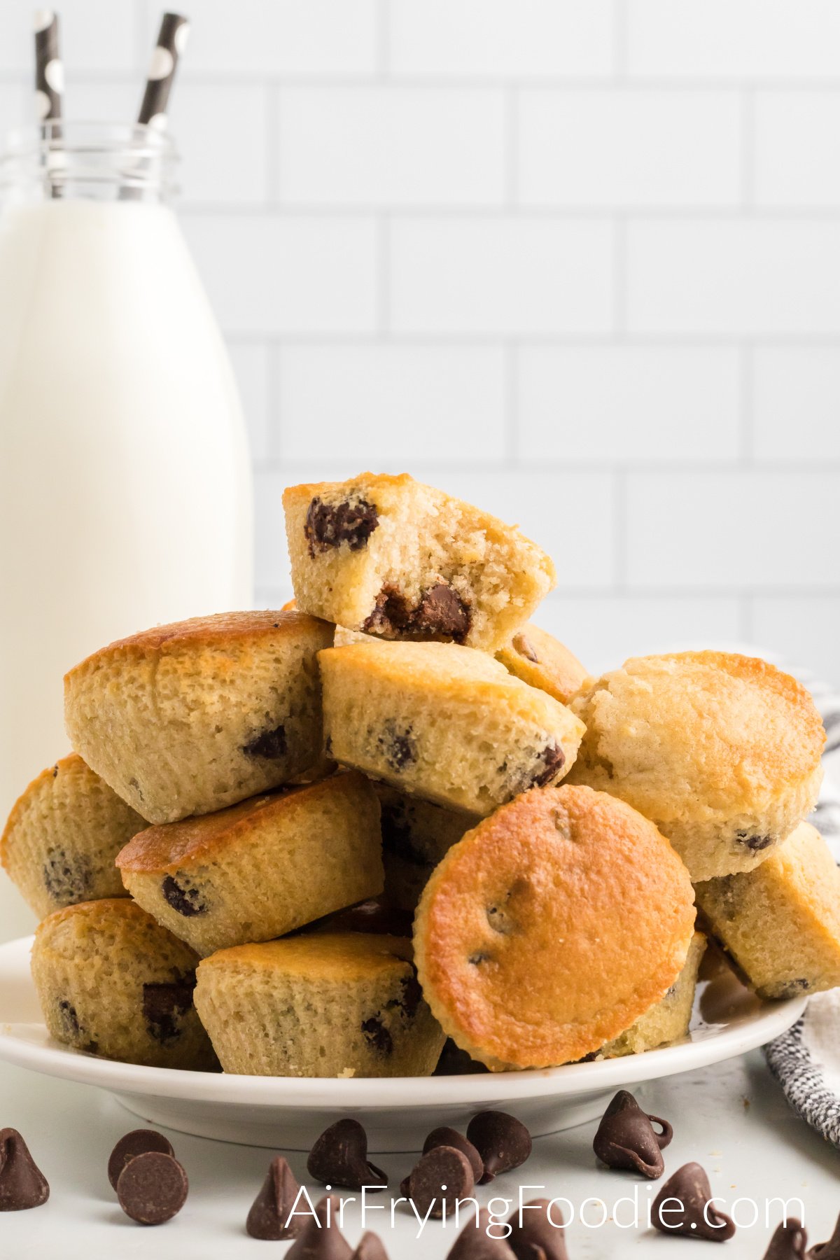 Chocolate chip muffins, after being cooked in the air fryer stacked on a round white plate with chocolate chips on the table in front of the plate and a tall glass bottle of milk with two straws. 