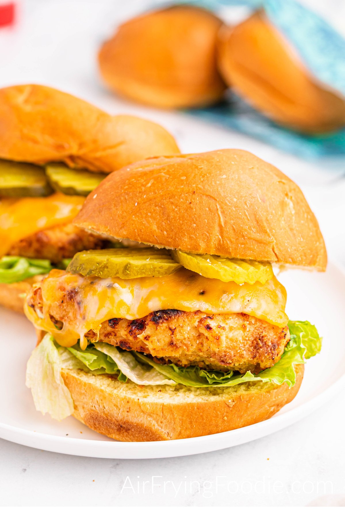 Two air fryer copycat chick oil a sandwich fully cooked and served on a white round plate dressed with pickles, cheese, chicken breast, lettuce, and chick oil sauce. Two extra buns halfway out of the bag are in the top right of the picture. 