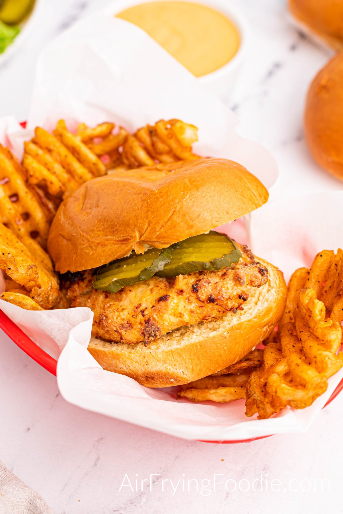 Chicken sandwich fully cooked from the air fryer served with waffle fries in a red basket. There is a white bowl of sauce above the basket and a small portion of extra buns in the image's top right.