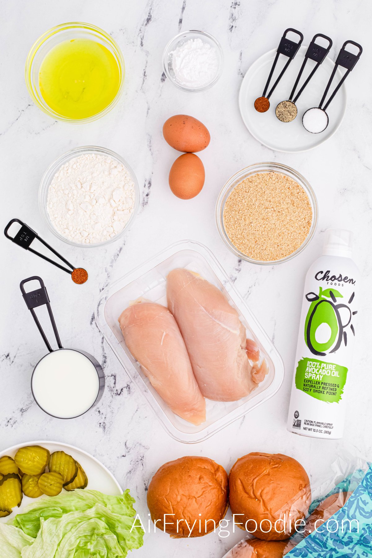 Picture of all the ingredients used to make copycat chick fil a sandwiches. Starting from the bottom of the photograph is an opened package of buns and a round white plate with lettuce and pickles. In the middle of the photo is a round black measuring cup of milk, a clear container of uncooked chicken, and a can of avocado oil. At the top of the image are four glass bowls, each containing a different ingredient; powdered sugar, flour, pickle juice, and breadcrumb. Two brown eggs, a round, and a white round plate with three measuring spoons, each holding a different ingredient; white pepper paprika and chili powder. 