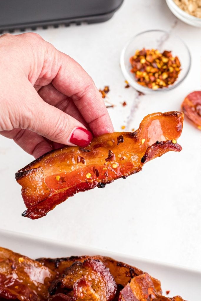 Juicy spicy bacon slices being held by a hand over the large plate. 