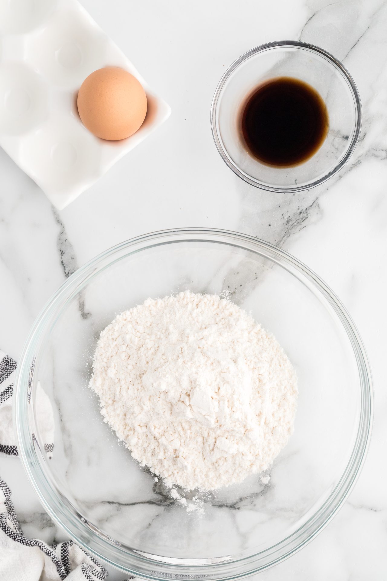 This picture shows a glass bowl with mixed flour, baking powder, and sugar. To the top left of the image is an egg container holding one brown egg and to the top right of the picture is a small bowl with vanilla extract.