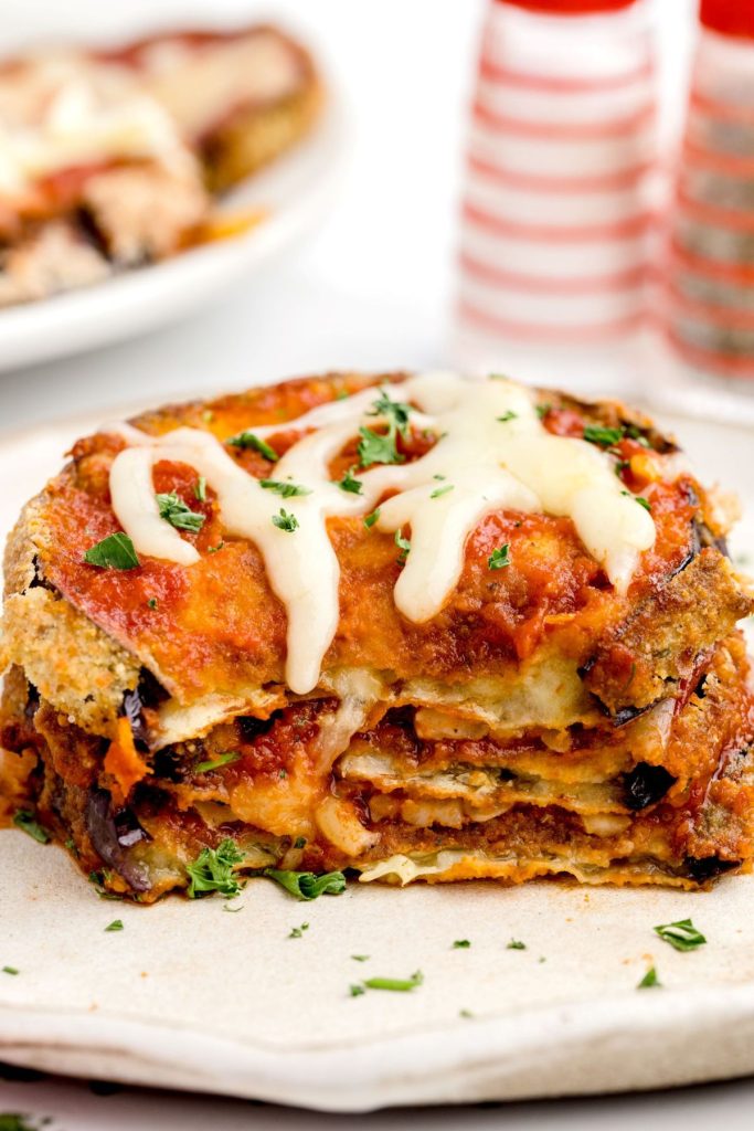 Crispy eggplant parmesan stacked on a white plate covered in marinara sauce and melted mozzarella cheese