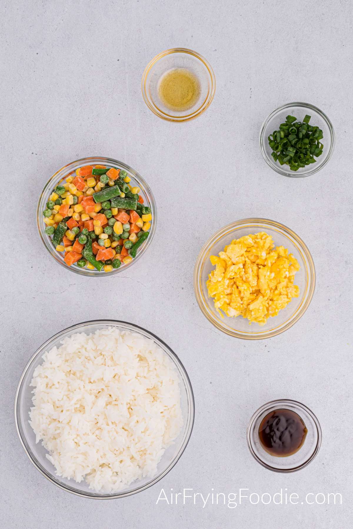 Overhead shot of the ingredients used to make air fryer fried rice. Three small glass bowls contain sesame oil, green onions, and oyster sauce. Two medium glass bowls have mixed vegetables, and the other includes eggs—one large bowl of white rice.