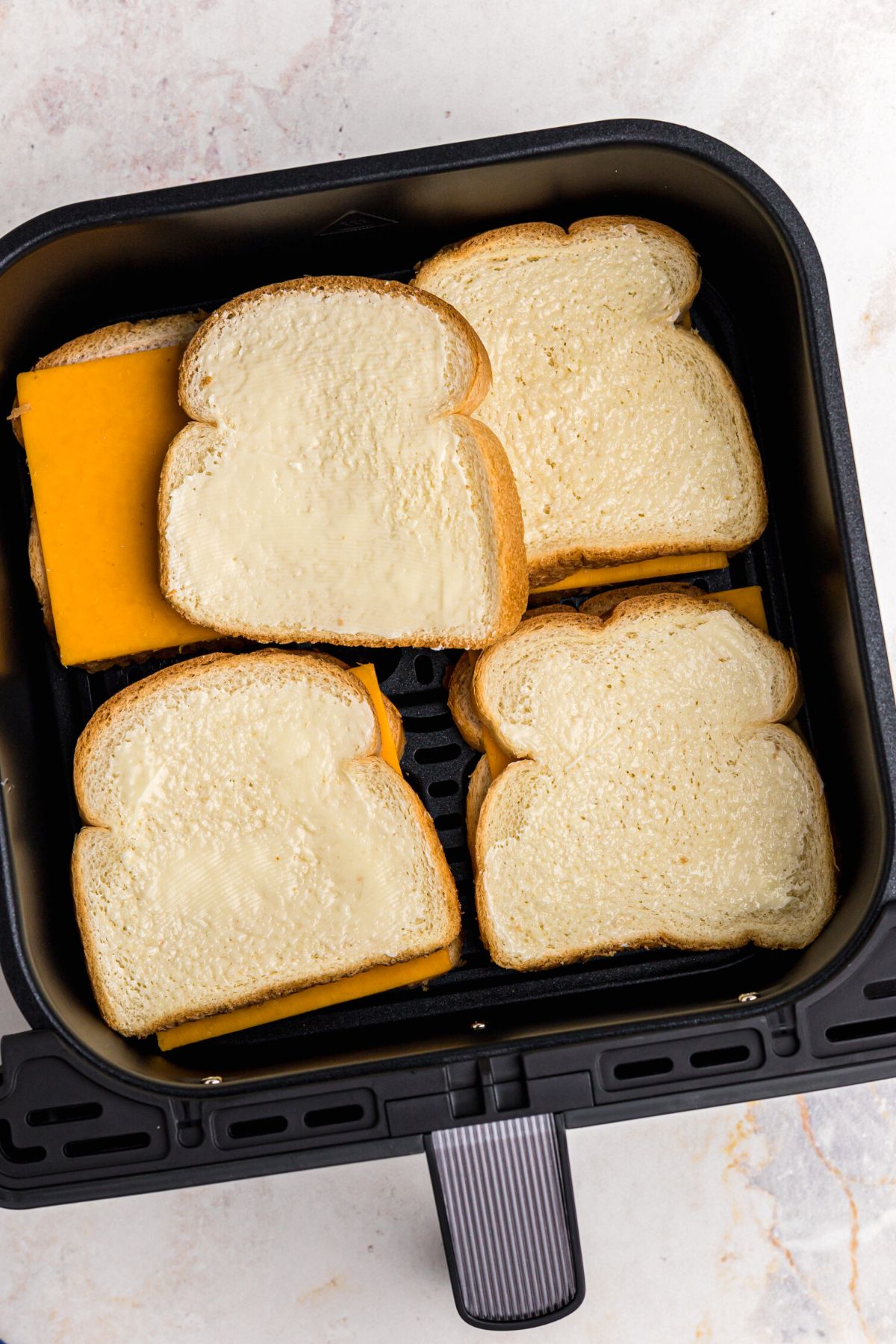 Bread slices with butter on one side, with a slice of cheese in the middle of bread slices, in the air fryer basket before being cooked.