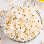 Clear glass bowl filled with fluffy popcorn and a small bowl of melted butter on a marble table.