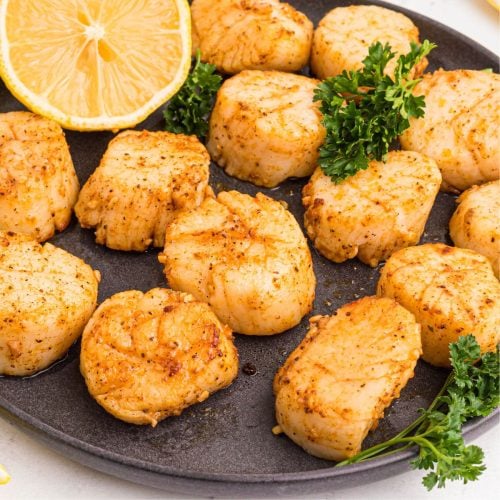 Golden juicy scallops on a dark plate seasoned and served with lemon wedges.