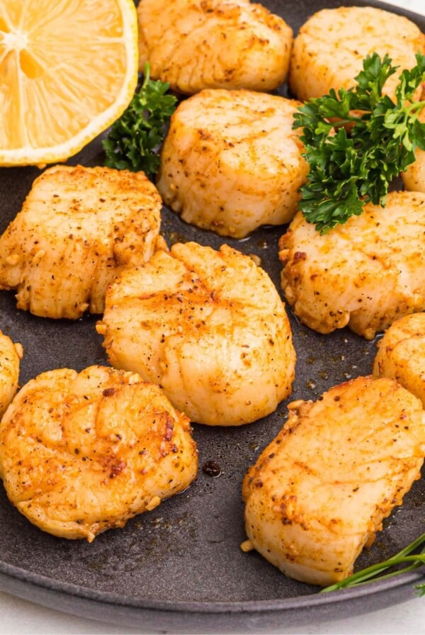 Golden juicy scallops on a dark plate seasoned and served with lemon wedges.