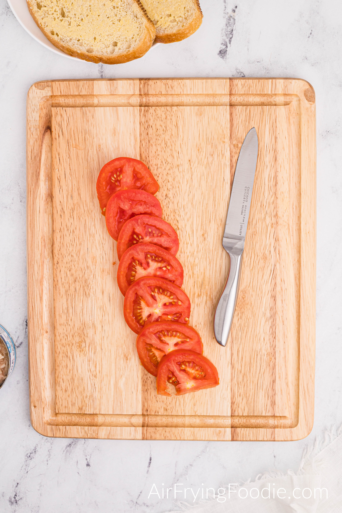 A tomato is sliced and lined on a cutting board with a butter knife on the right of the cutting board and two slices of Texas toast on a round white plate above the cutting board.