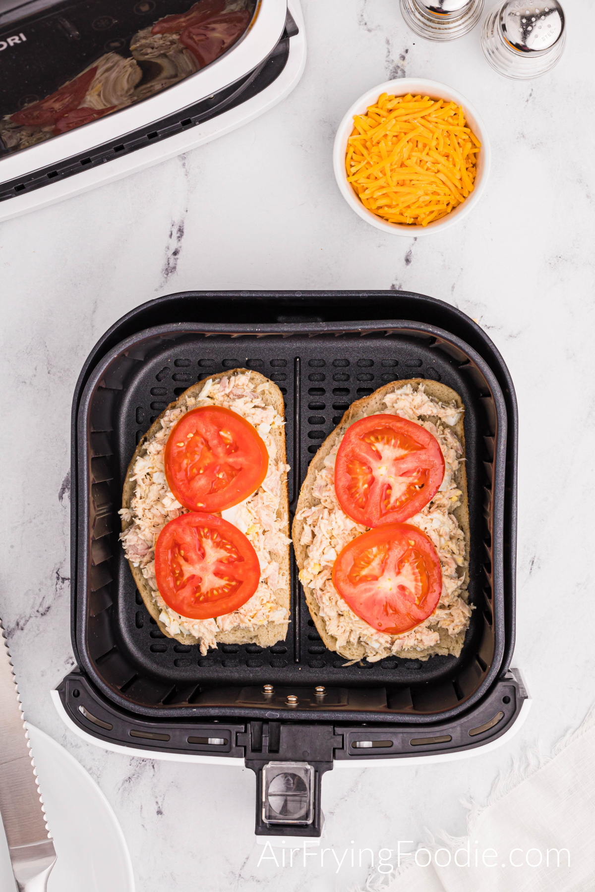 Two slices of Texas toast in the air fryer basket topped with tuna and two slices of tomato on each piece of Texas toast. There is a small bowl of shredded cheese, a salt, and pepper shaker to the top right of the air fryer basket, and a small portion of the air fryer base if at the top left.