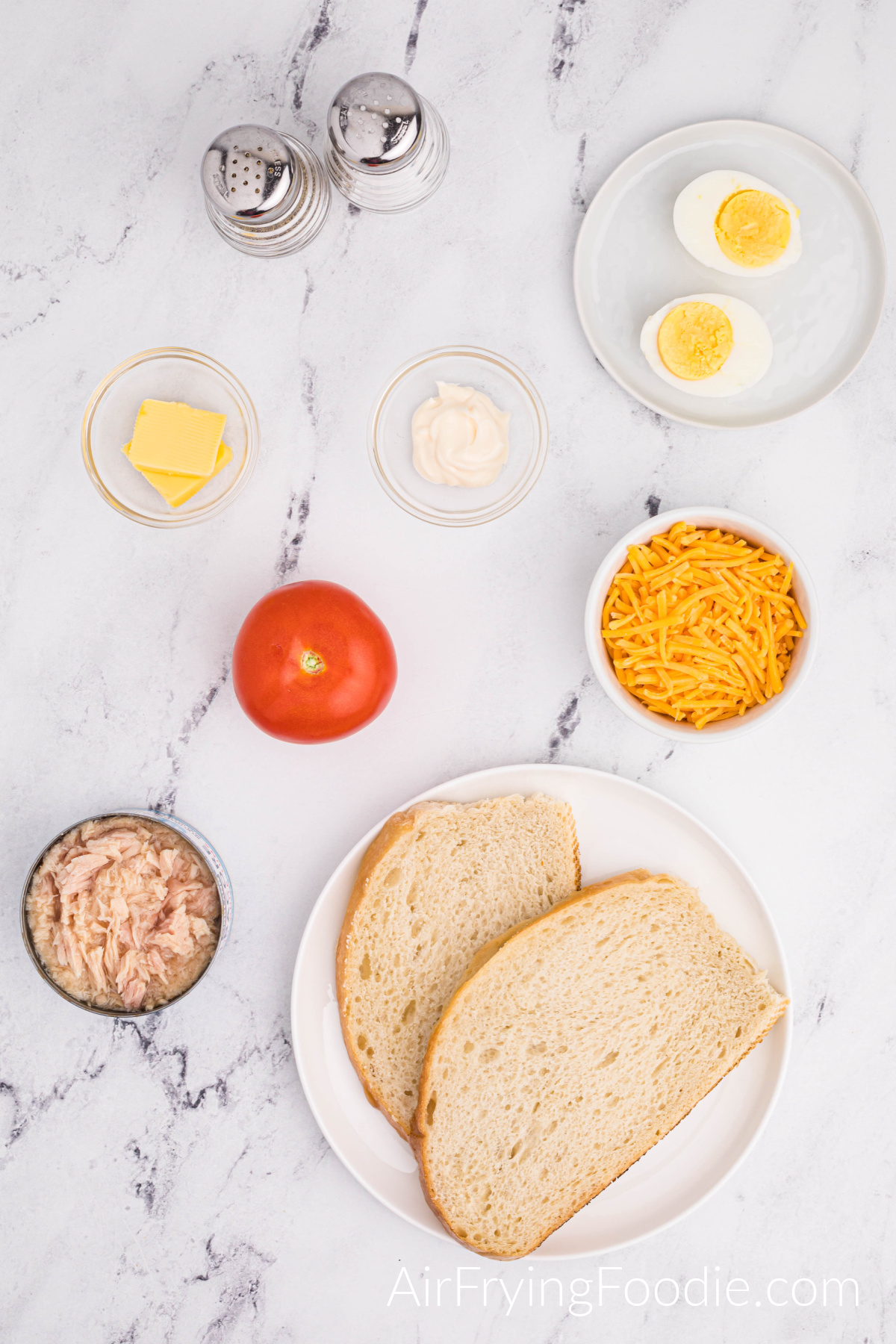 Overhead shot of the ingredients used to make a tuna melt in the air fryer. Two round white plates; one with two slices of Texas toast and the second with one egg boiled and cut in half. Three small glass bowls contain tuna; the other has butter and mayonnaise—a small white bowl of shredded cheese, one whole tomato, and a salt and pepper shaker.