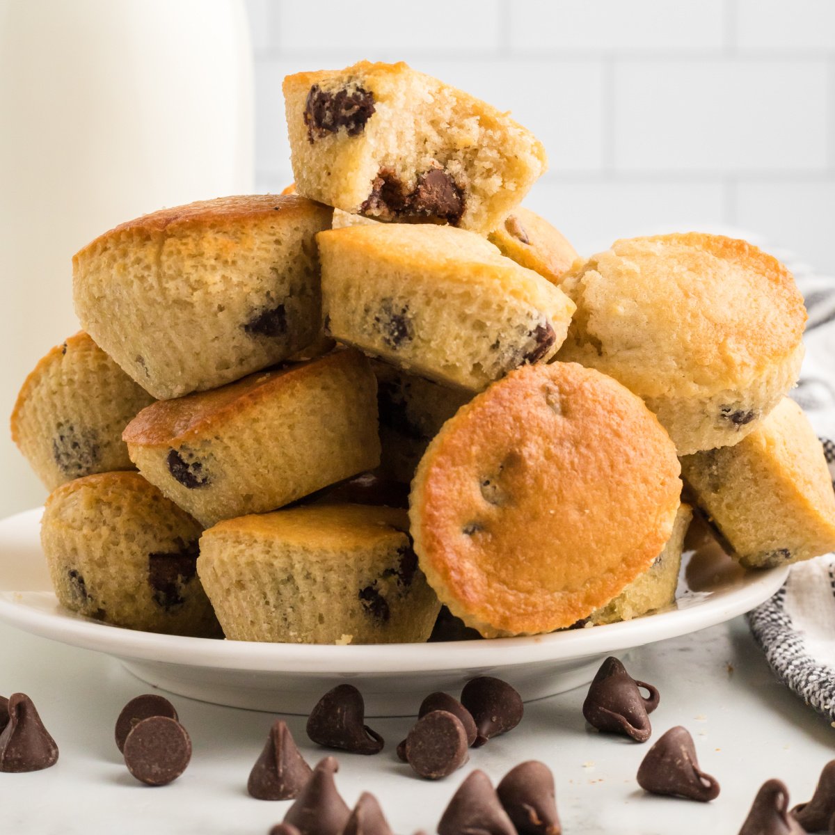 Round white plate fully stacked with chocolate chip air fryer muffins baked in the air fryer with chocolate chips sprinkled around on the table in front of the plate.