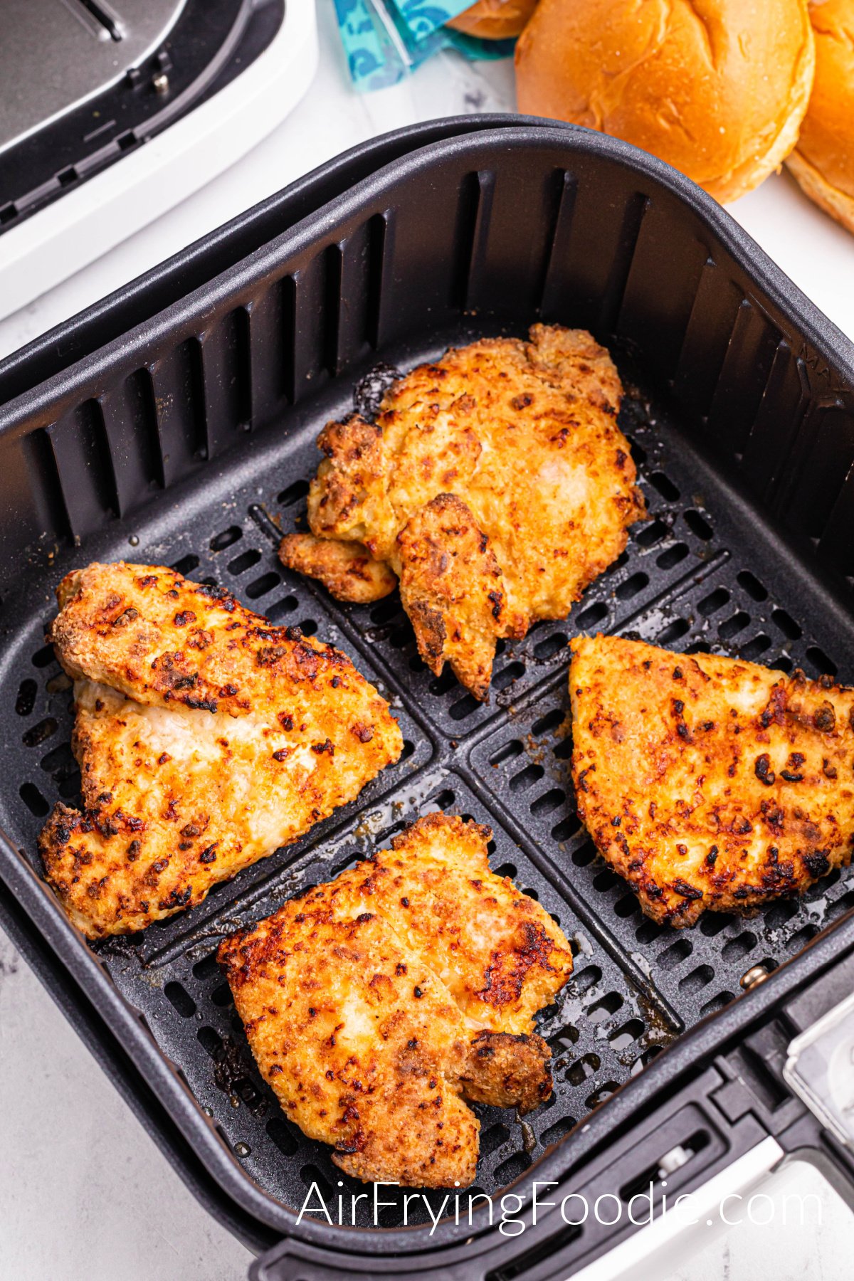 A picture of four pieces of chicken halfway cooked in the air fryer basket before flipping over. The hamburger buns are at the top right corner of the picture.