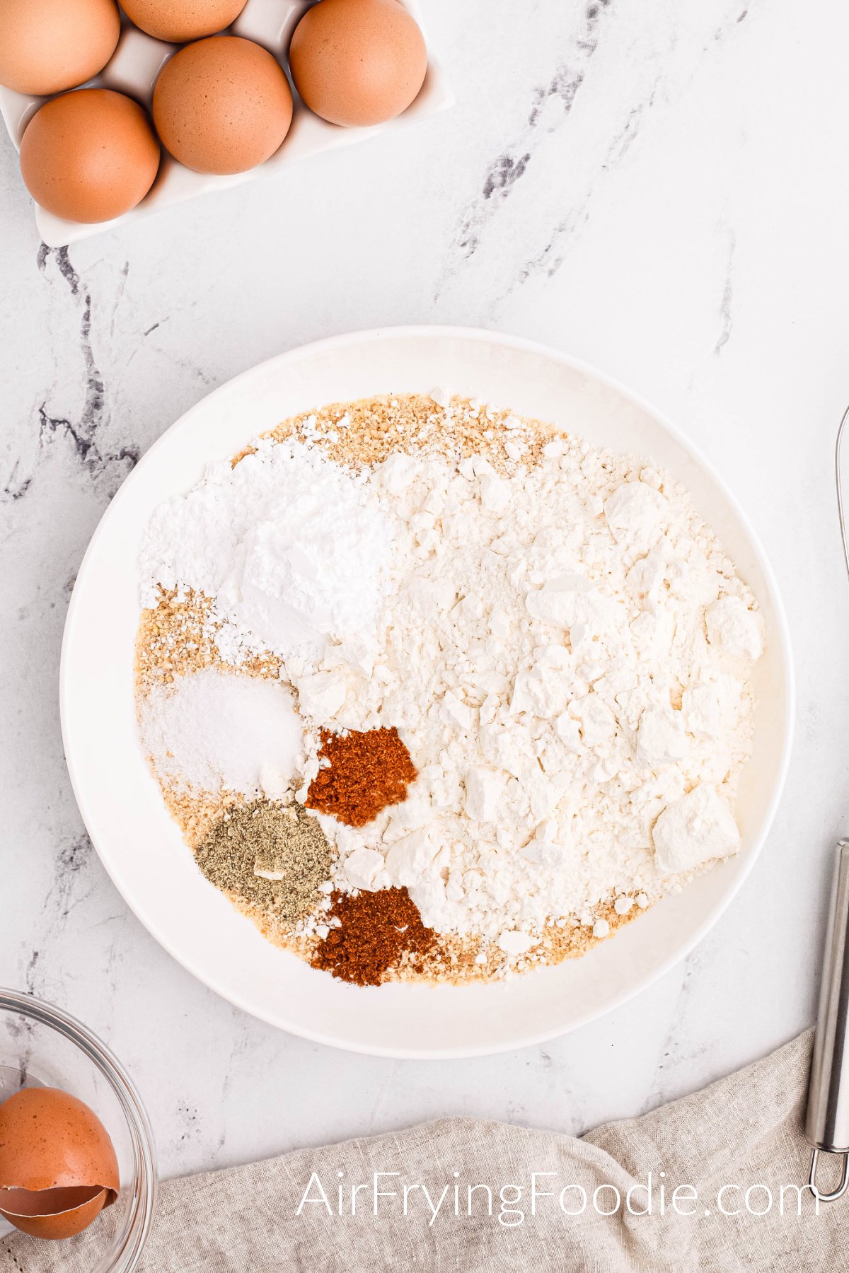 Round white bowl with all the dry ingredients poured in; breadcrumbs, flour, powdered sugar, salt, paprika, pepper, and chili powder.